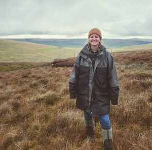 An image of WRT Volunteer and Engagement Officer, Jade Harley, wrapped up in winter clothing on rough grassland with rolling hills in the background.